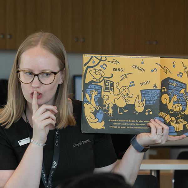 Children's Librarian reading a book and making a shush sign.