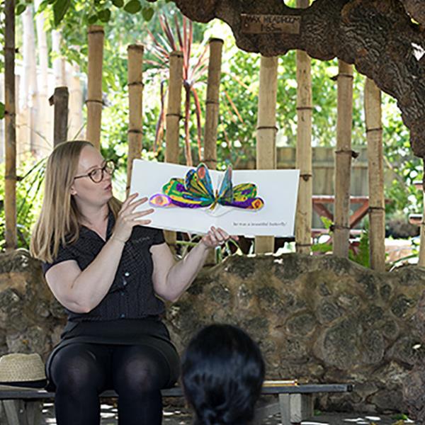 Female presenter reading a picture book to audience under a tree