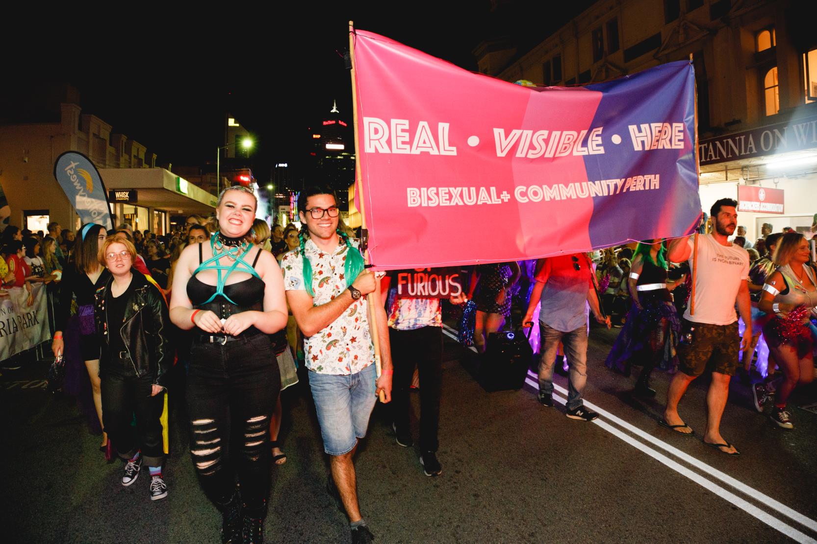 Pride parade participants holding sign that says 'Real. Visible. Here. Bisexual+ Community Perth'