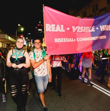 Pride parade participants holding sign that says 'Real. Visible. Here. Bisexual+ Community Perth'