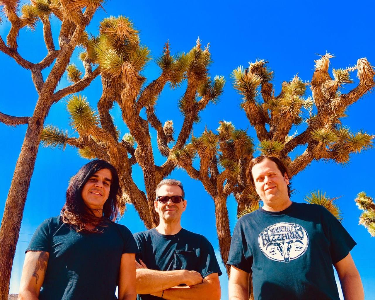 Three band members in black shirts with bright blue skies and trees behind them