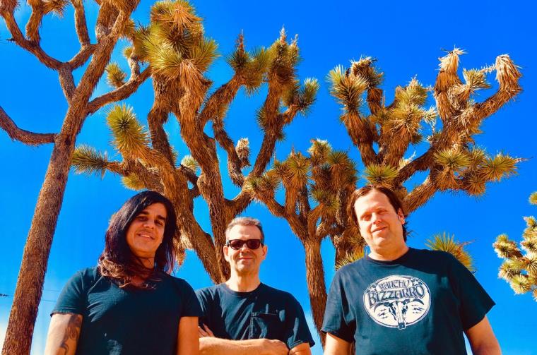 Three band members in black shirts with bright blue skies and trees behind them