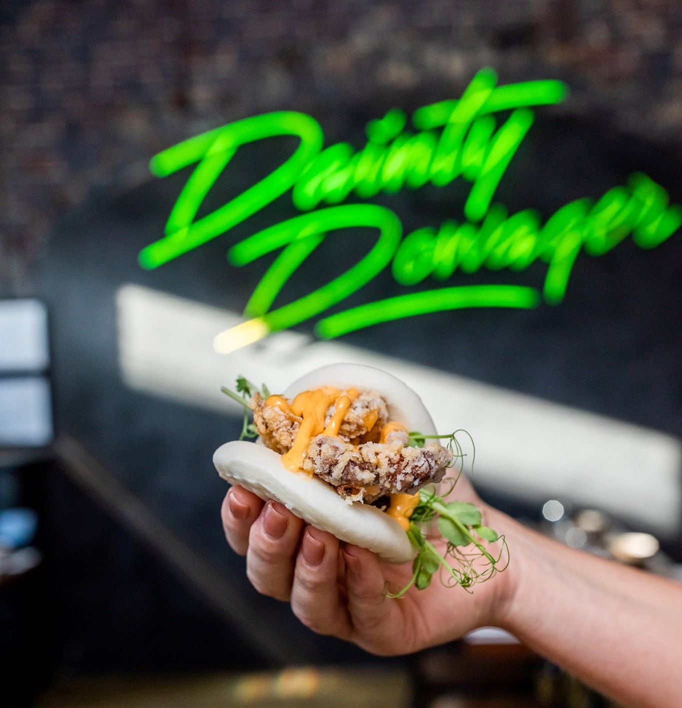 Hand holding a bao with a green neon sign in background 'Dainty Dowager'