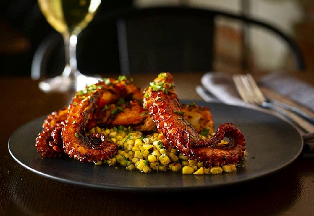Plate with octopus and corn dish in foreground and glass of white wine in background