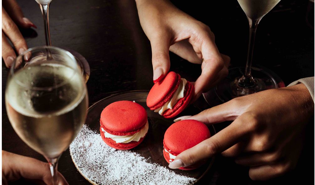  	 Glasses of champagne and hands reaching for red macaroons
